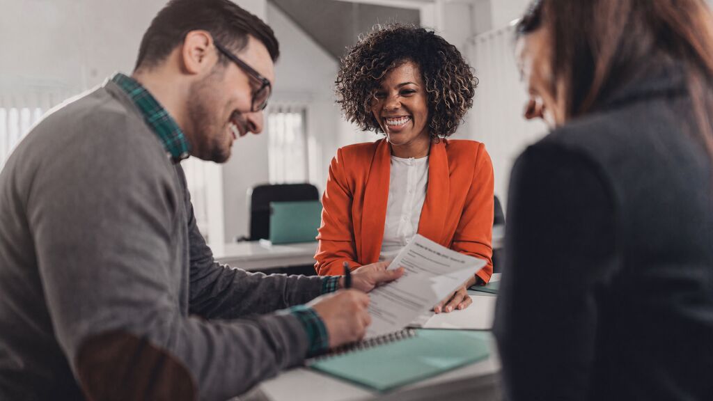 Smiling financial advisor in an orange blazer reviewing paperwork with a couple signing documents across a desk, depicting client onboarding and loan or insurance processes that UiPath agentic automation and AI agents accelerate.