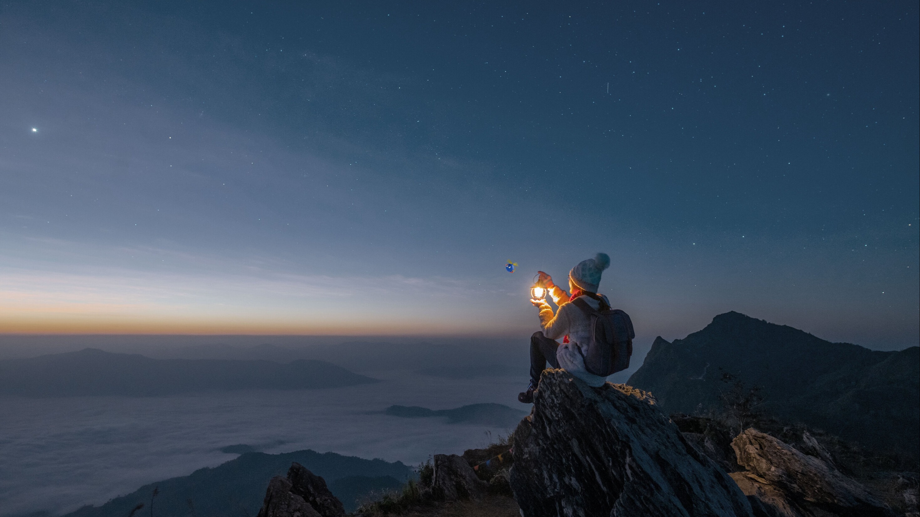 A person holding a lamp, sitting on the top of a cliff gasping upon a sunset or sunrise. 
