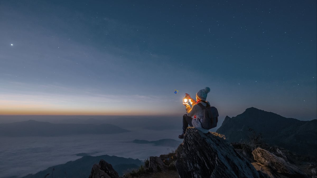 A person holding a lamp, sitting on the top of a cliff gasping upon a sunset or sunrise. 