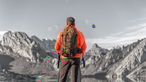 A person in an orange jacket with a backpack stands on a rocky landscape, gazing at a mountain range and a small UiPath bot flying in the sky.