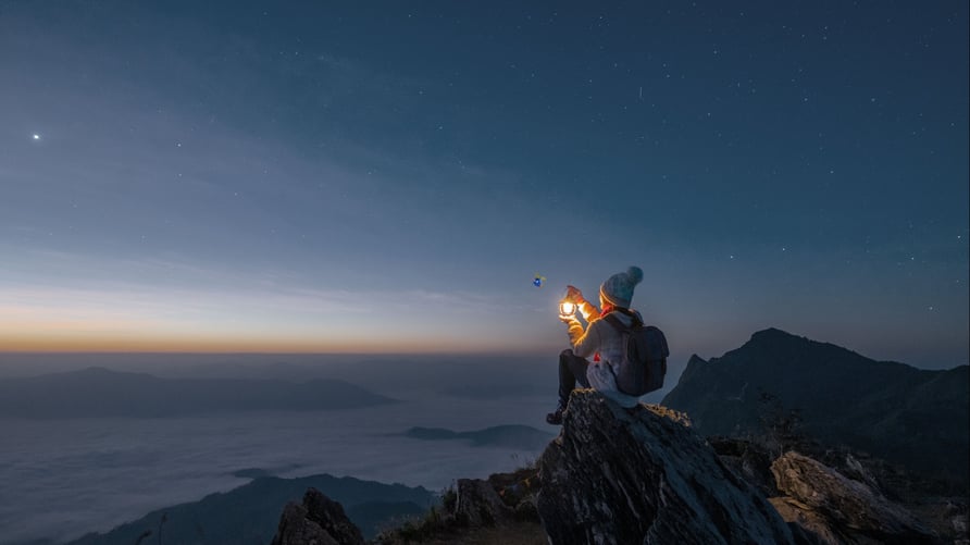 A person holding a lamp, sitting on the top of a cliff gasping upon a sunset or sunrise. 